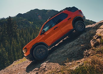 Orange off-road SUV climbing a steep rocky hill with mountains and trees in the background.